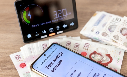 A photo of a in-home display of a smart meter, bundles of £10 and £50 notes and a phone displaying a clients energy account.