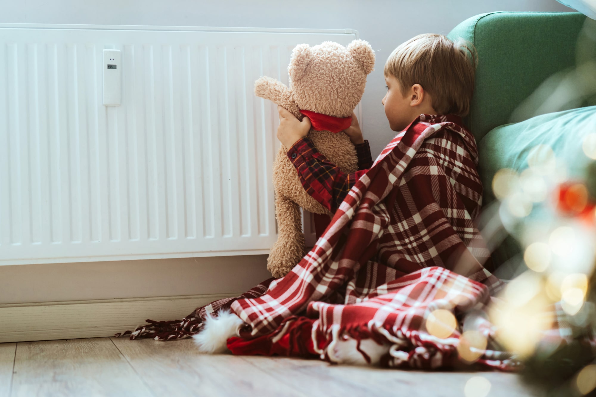A photo of a child living in fuel poverty. He has a large blanket over him and he is playing with a teddy bear near a radiator.