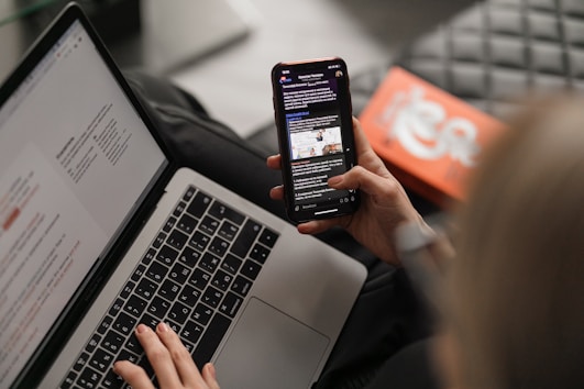 A photo of a woman looking at information on a laptop and holding a mobile phone in the other hand.