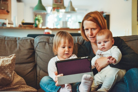 A photo of a mother with two children sat on her knee. They are all looking at a tablet.