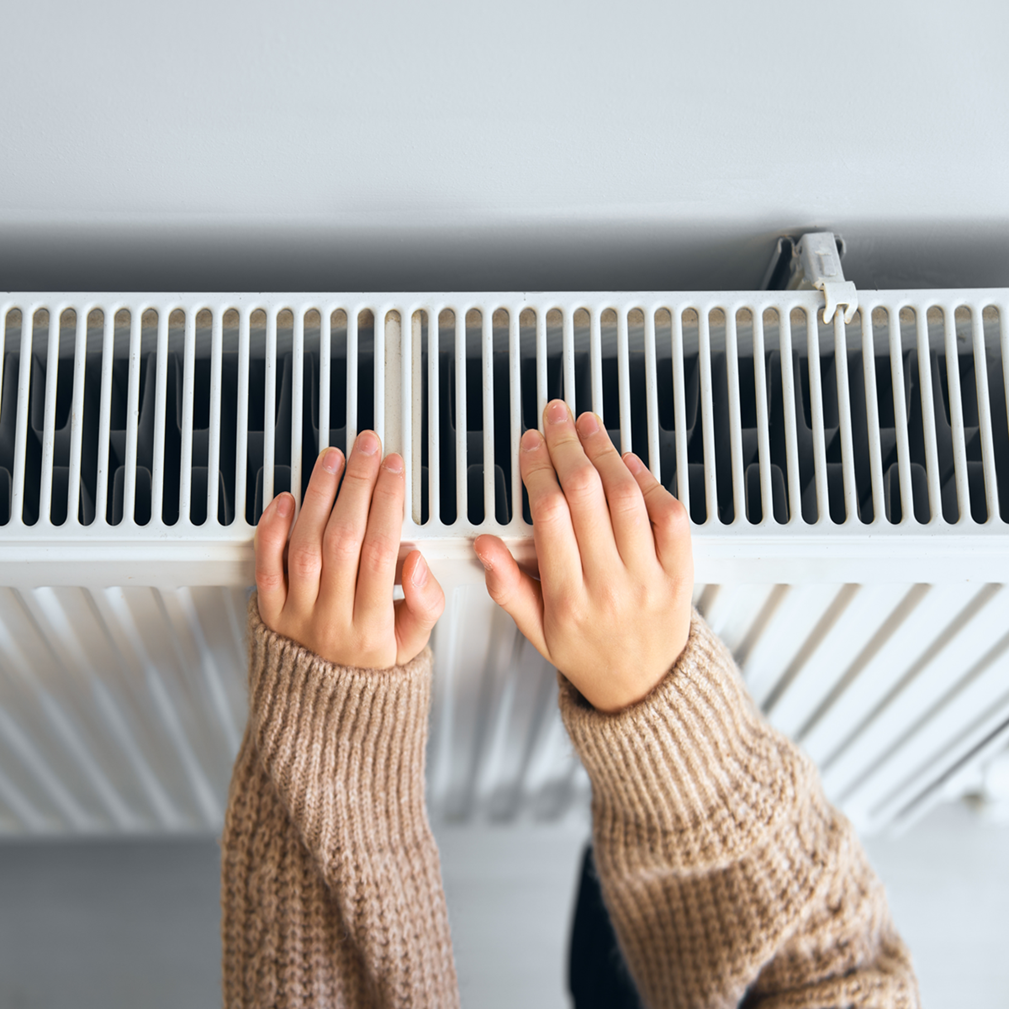 Photo of two hands held above a radiator. The person in a beige jumper is trying to keep warm.