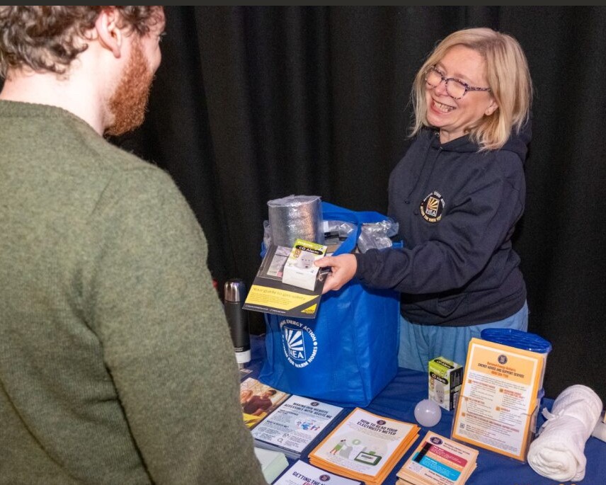 National Energy Action adviser gives support to a client. She is handing out a Winter Warmth Support Pack at a community event which had a table covered in advice leaflets.