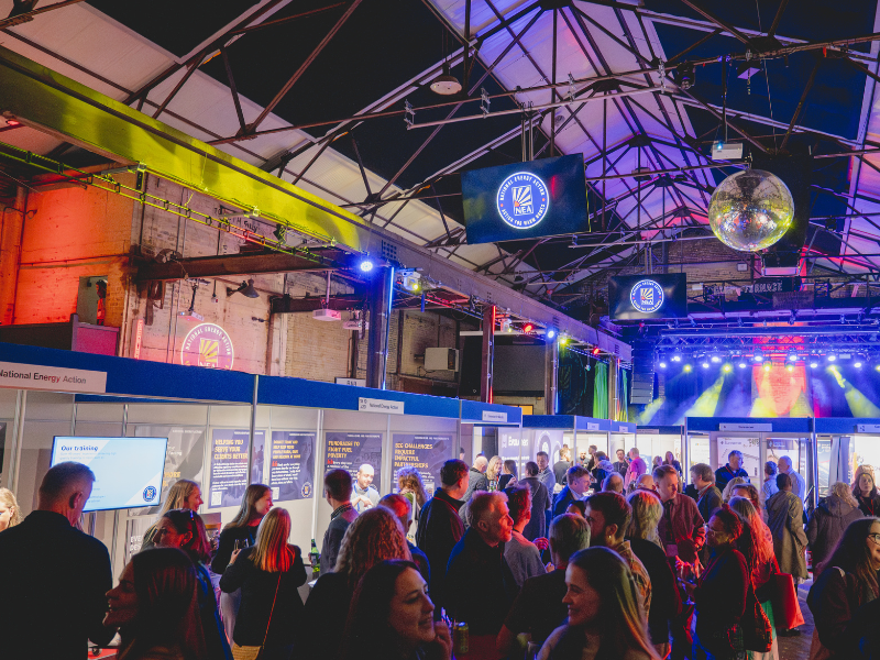 A photo of the exhibition at National Energy Action's fuel poverty conference. Delegates are gathered and are networking in a large space with stands and the NEA logo displayed on the ceiling.