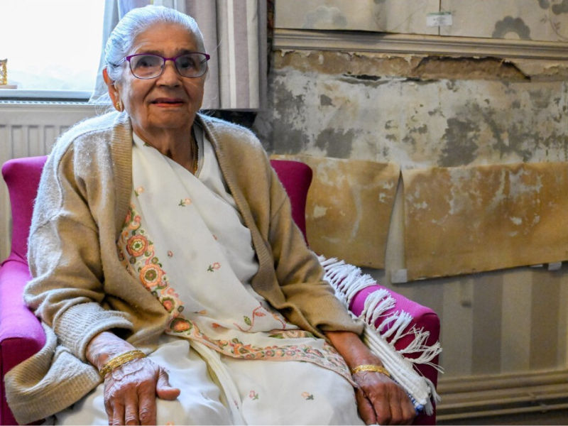 A photo of a woman sitting in an armchair in warm clothing. Her house is covered in damp and mould.