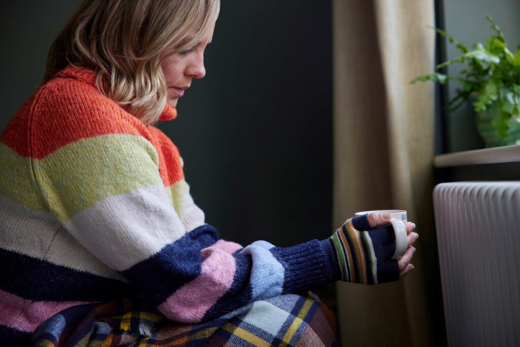 A woman wearing warm clothes and gloves inside. She is holding a warm drink near a radiator. 