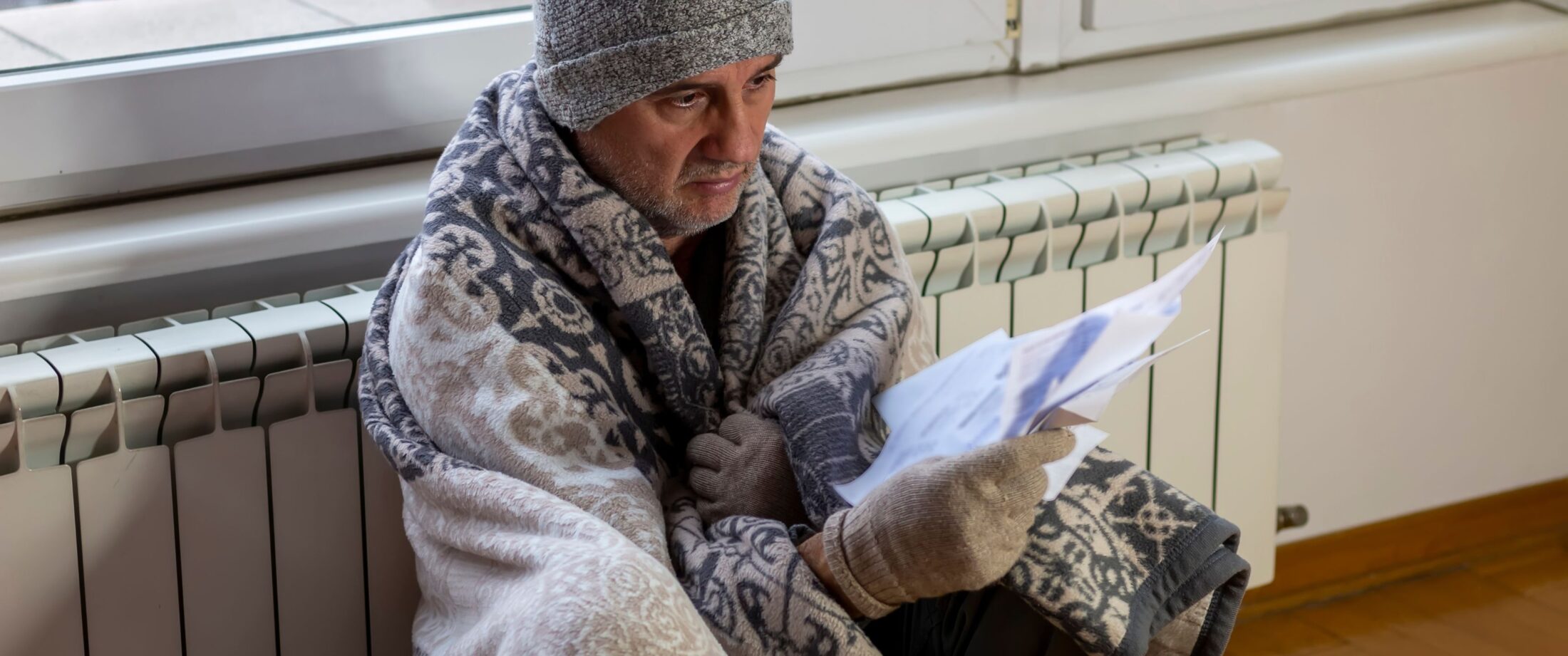 A man in a blanket and beanie sits on the floor by a radiator, looking at a bill