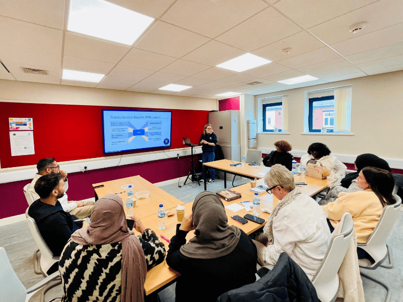 Seven adults at a community advice session. They are listening to an Adviser from National Energy Action giving a presentation on how to save money on their energy bills.