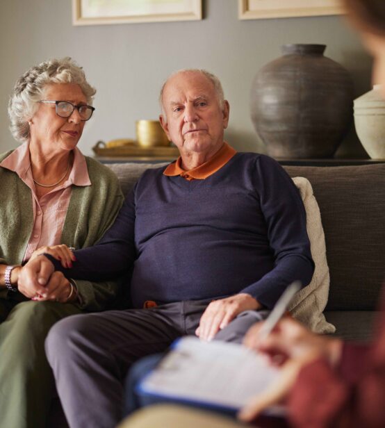 Stock photo of an elderly couple sitting on a sofa together holding hands. They are speaking to someone sitting on a chair in their living room with clipboard.