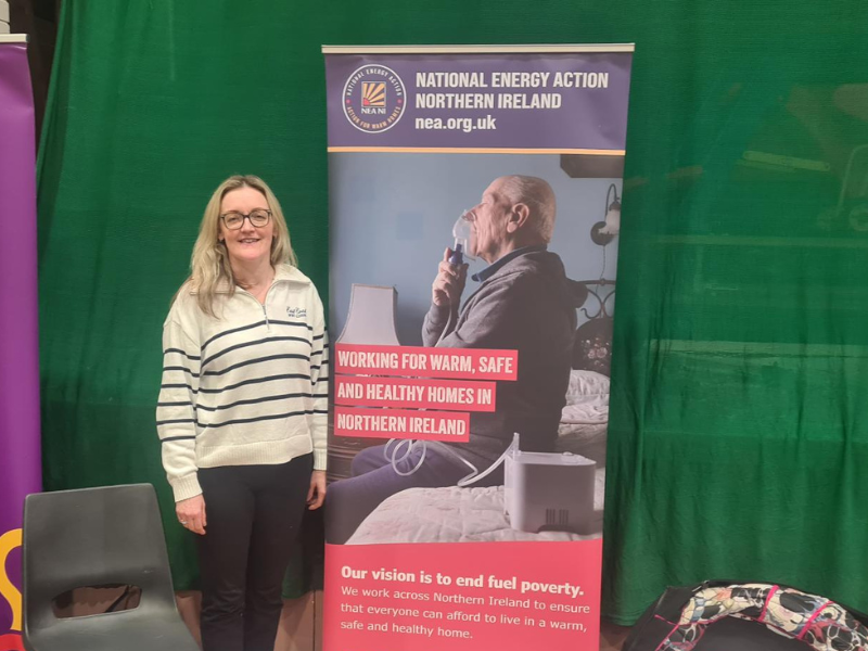 A photo of National Energy Action's NI Training Officer stood in front of a banner that says 'Working for Warm, Safe and Healthy Homes in Northern Ireland.'