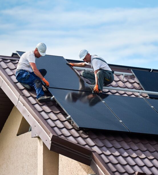 A photo of two workers on a roof installing solar panels on a bright day with a blue sky with some clouds.