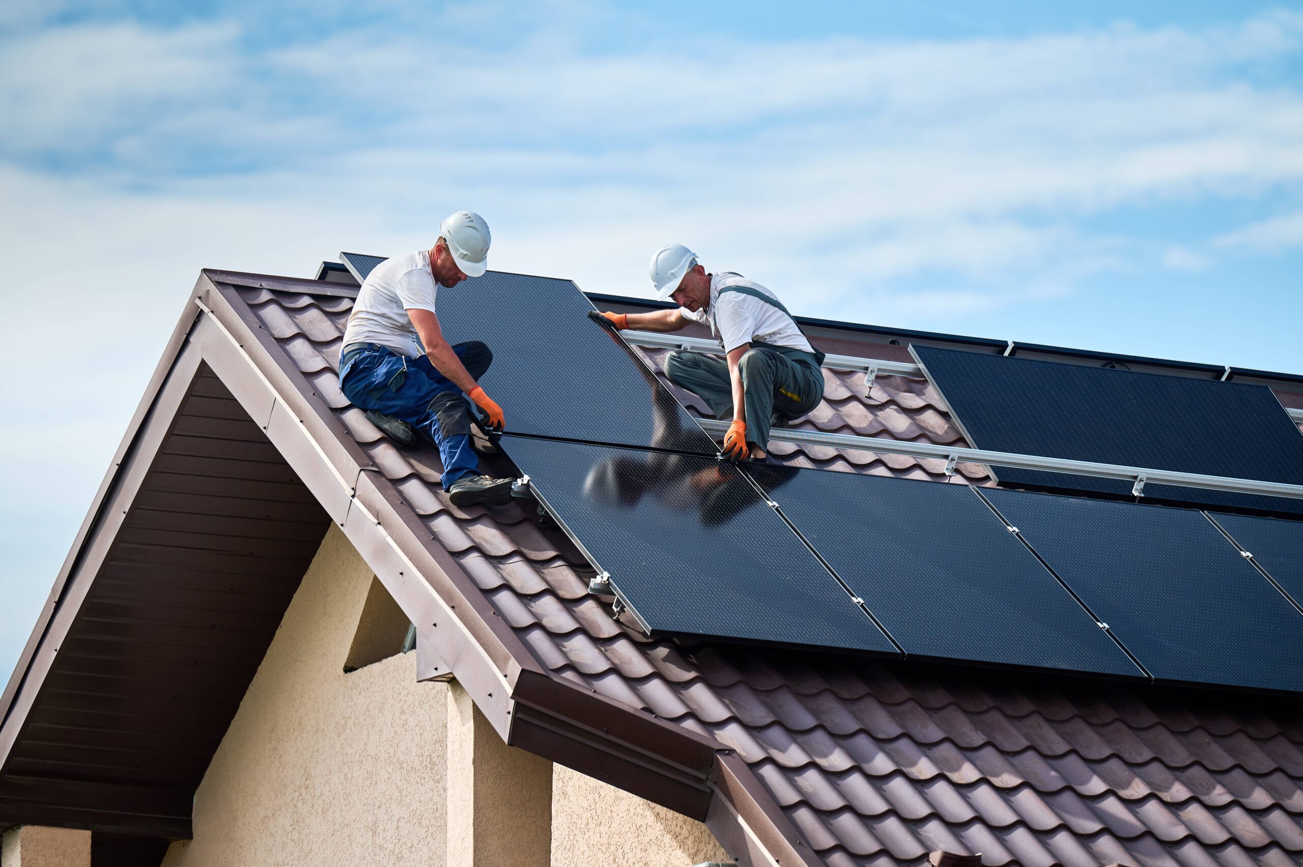 A photo of two workers on a roof installing solar panels on a bright day with a blue sky with some clouds.