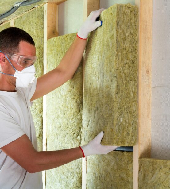 Stock photo of a worker installing insulation in a wall. He is wearing safety goggles, a mask and gloves.