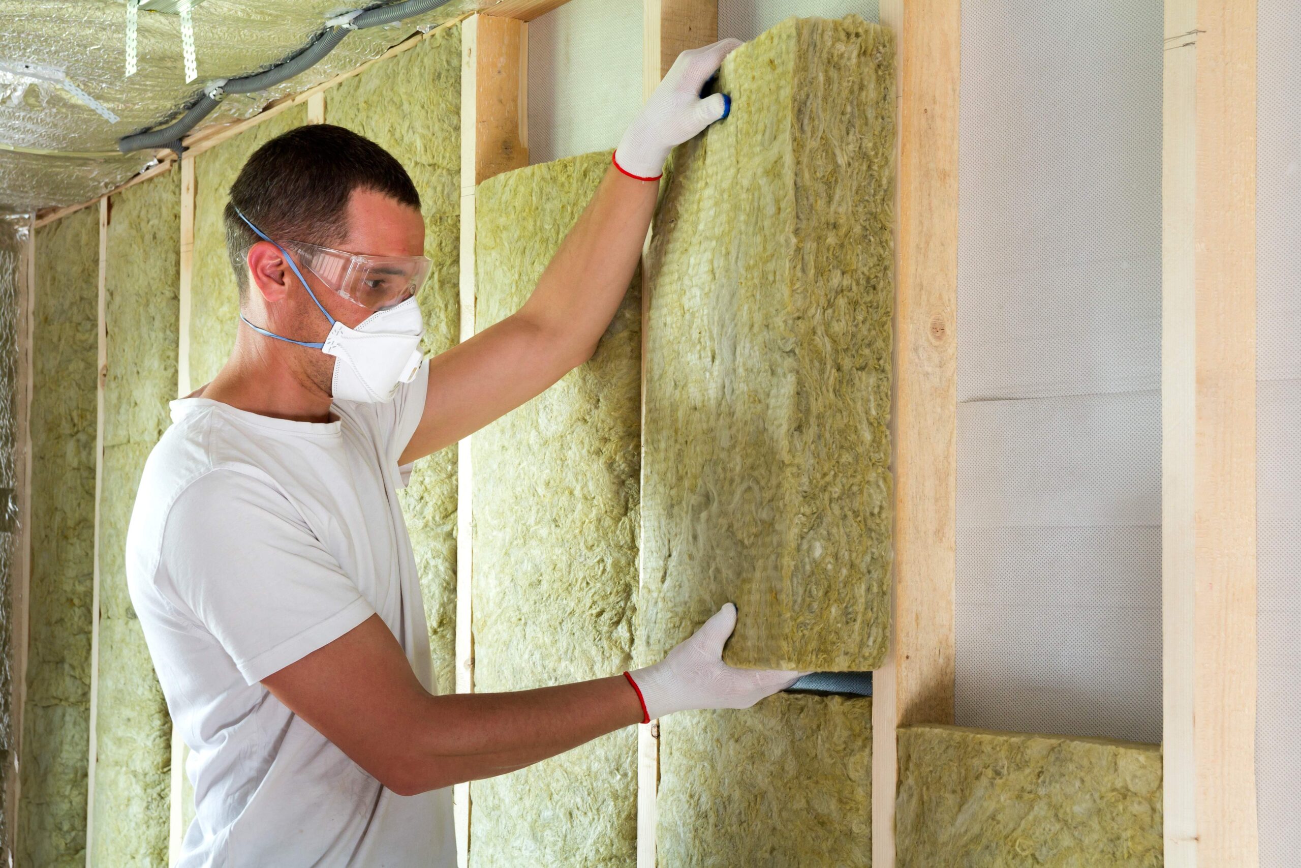 Stock photo of a worker installing insulation in a wall. He is wearing safety goggles, a mask and gloves.