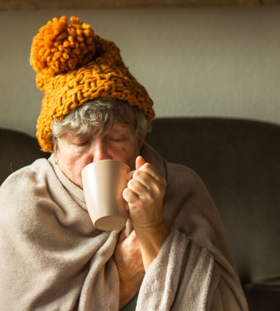 A older woman wearing a wooly hat and wrapped in a large blanket. She is drinking from a mug.