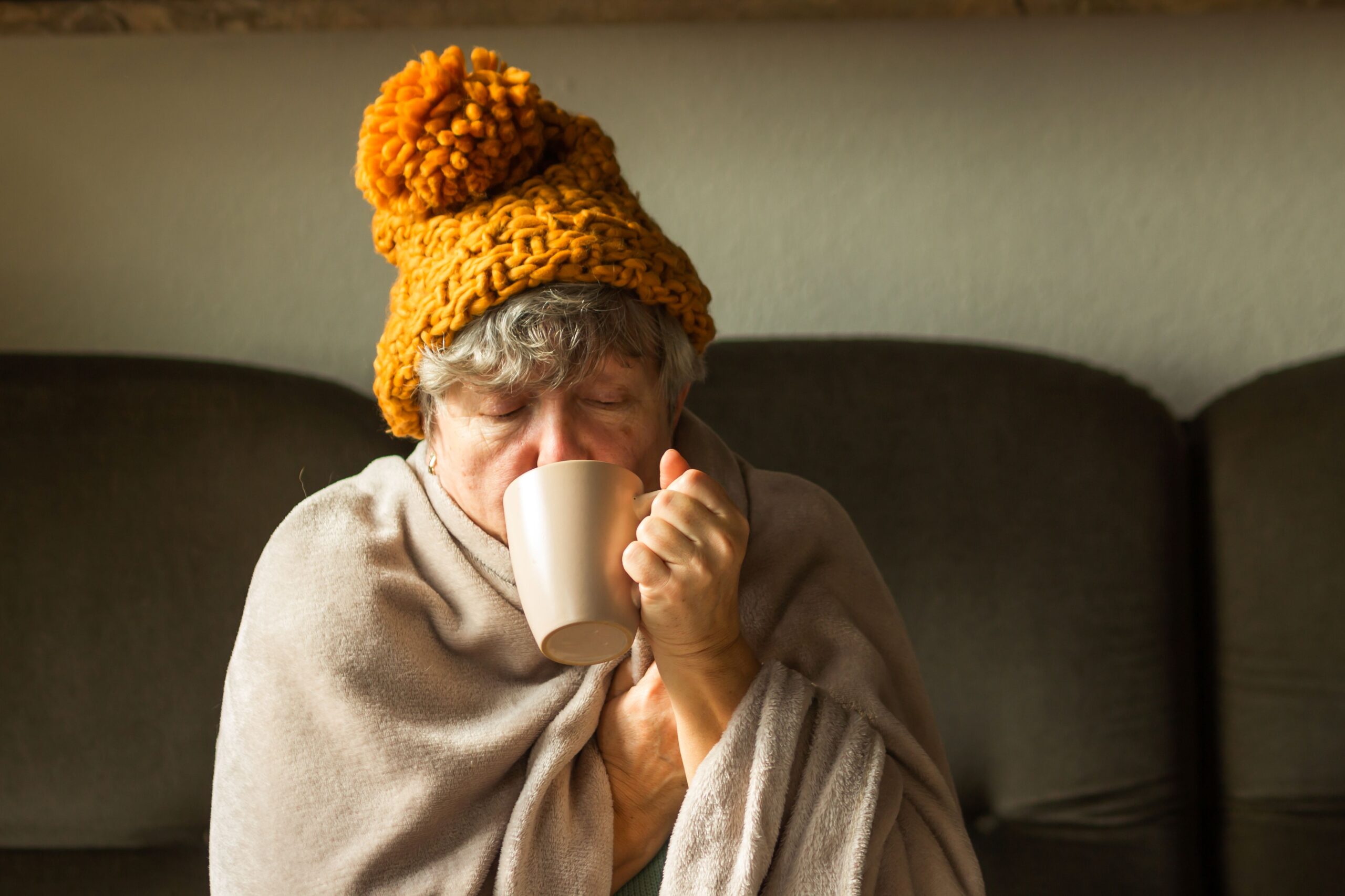 A older woman wearing a wooly hat and wrapped in a large blanket. She is drinking from a mug.