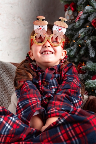 A photo of a child looking happy at Christmas. He is wearing novelty christmas glasses and is sat on the sofa with a Christmas tree in the background.