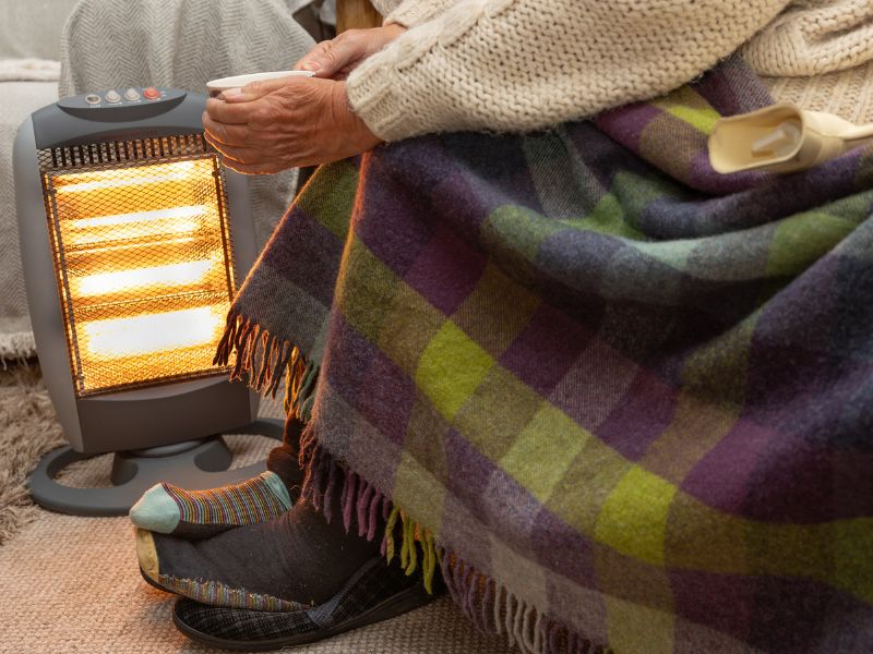 An elderly person in a knitted sweater and checkered blanket warms their hands near a glowing space heater, creating a cozy and comforting atmosphere.