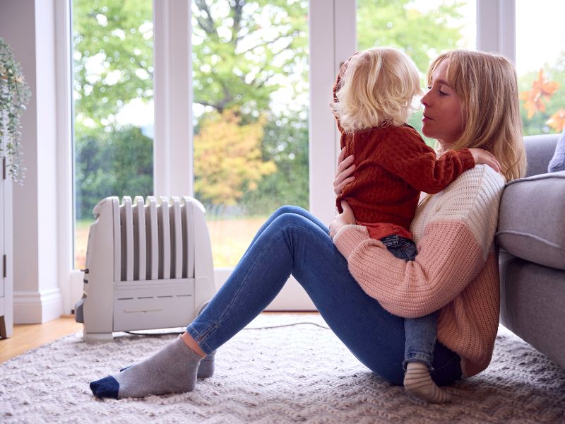Mother sitting on a carpeted floor holding a young child beside an electric heater in a bright living room, both dressed in warm knit clothing