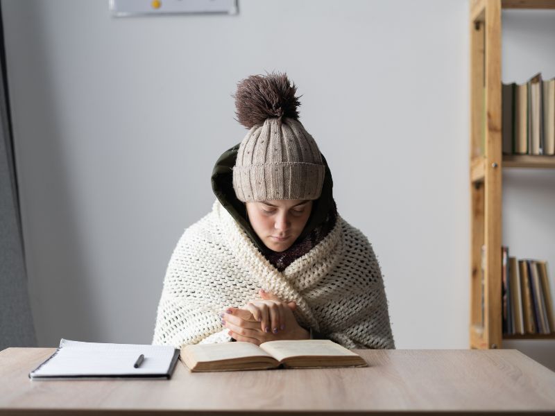 Person in a knitted hat and blanket reading a book at a table