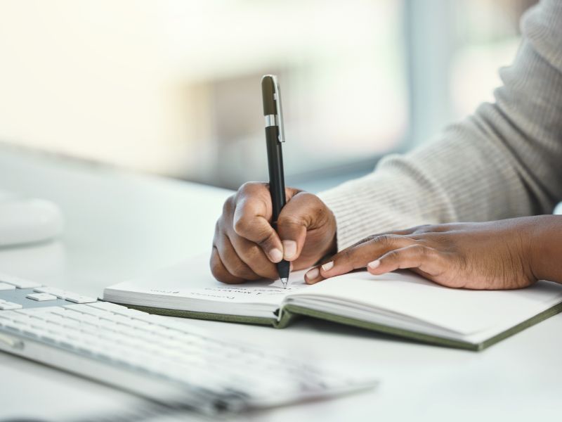 A person writing in a notebook with a pen on a desk with a keyboard nearby