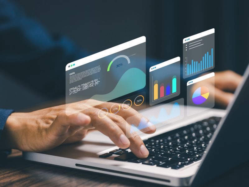 Hands interact with a laptop displaying floating analytics dashboards, bar and pie charts, and performance metrics in a modern, data-driven scene.